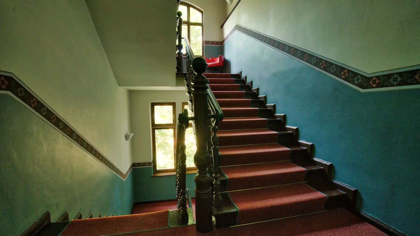 Altbau-Treppenhaus mit Holzgeländer, roten Stufen und Tageslicht